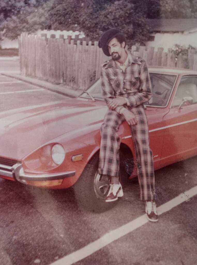 A person wearing a plaid outfit and a beret sits on the hood of a vintage red sports car in a parking lot. A wooden fence and trees are visible in the background.