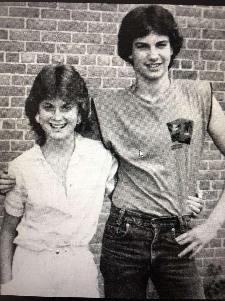 A young man and woman stand next to each other smiling in front of a brick wall. The man has his arm around the woman's shoulders. The woman is wearing a light-colored jumpsuit, and the man is wearing a sleeveless shirt and jeans.