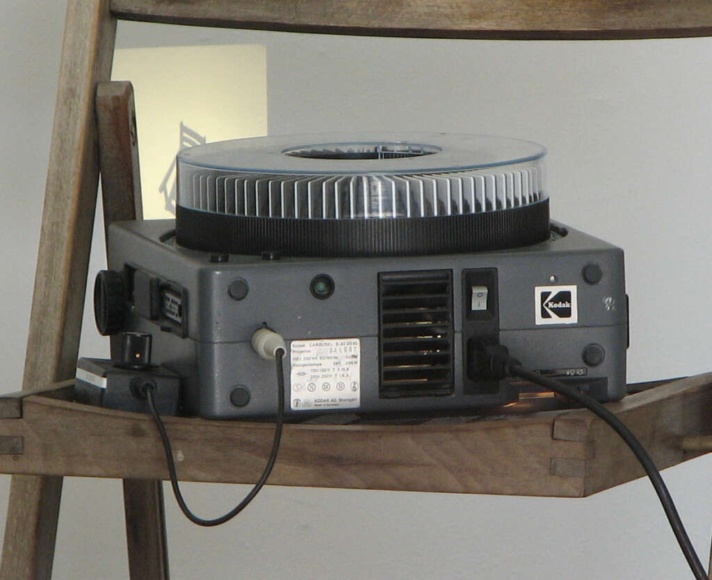 A gray Kodak carousel slide projector sits on a wooden shelf. The projector has a circular tray loaded with slides, and various connectors plugged into the back. Its classic design and slightly worn appearance hint at frequent use.