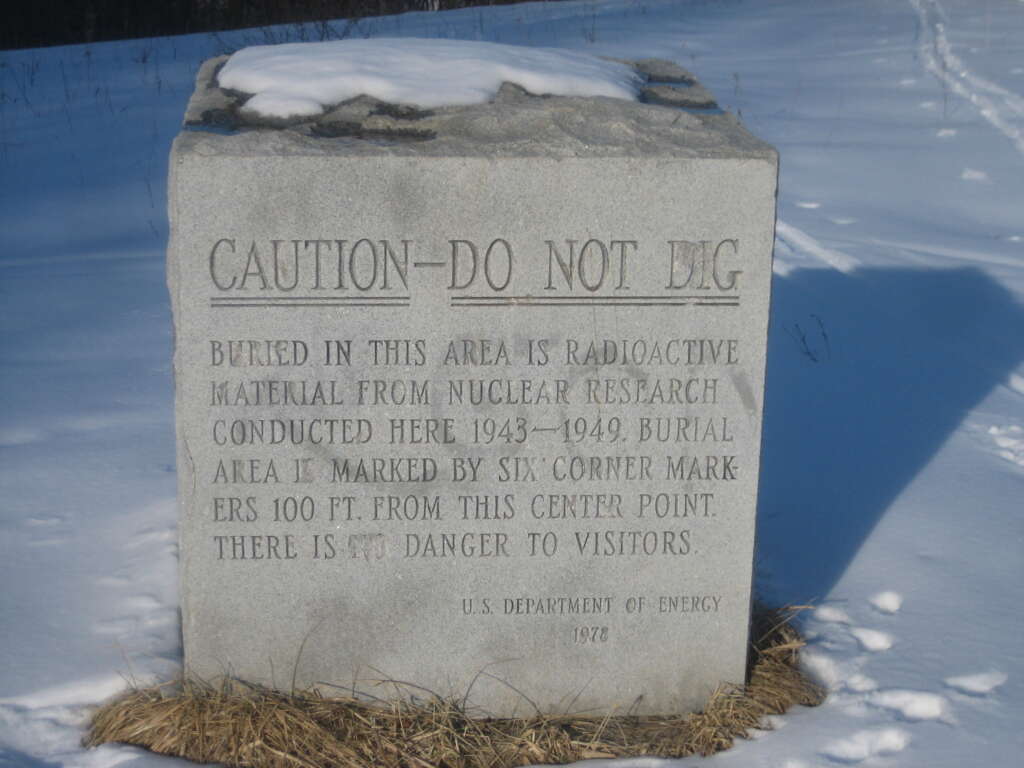 A snow-covered, weathered stone marker in a field, displaying an engraved warning that reads: "CAUTION—DO NOT DIG. Buried in this area is radioactive material from nuclear research conducted here 1943–1949. Burial area is marked by six corner markers 100 ft. from this center point. There is no danger to visitors. U.S. Department of Energy 1978.