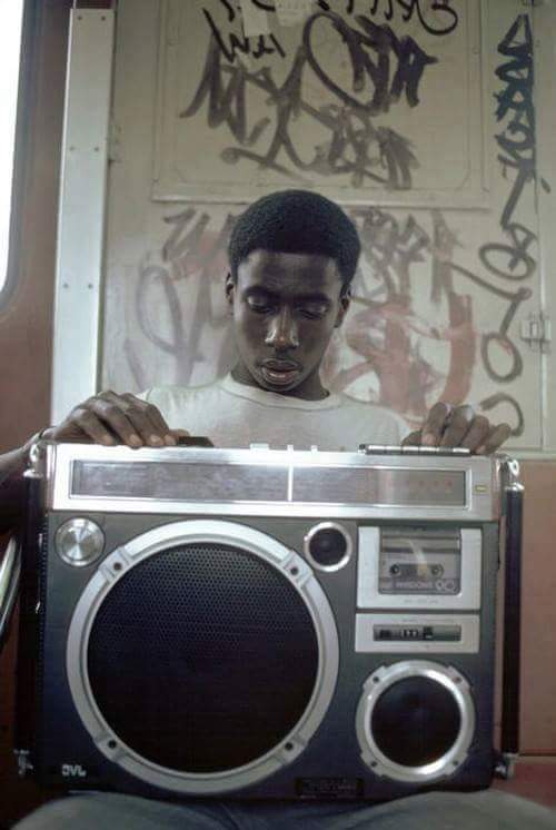 A young man sits on a train, focusing intently on a large boombox he is holding. The background shows a wall covered in graffiti, and the atmosphere suggests a 1980s setting. The picture captures a moment of concentration and urban culture.