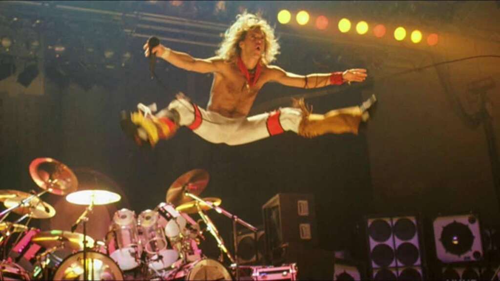 A rock singer is captured mid-air performing a high jump on stage, holding a microphone, with legs spread wide apart. The stage is set with drums and several speakers in the background, and colorful lights illuminate the scene from above.