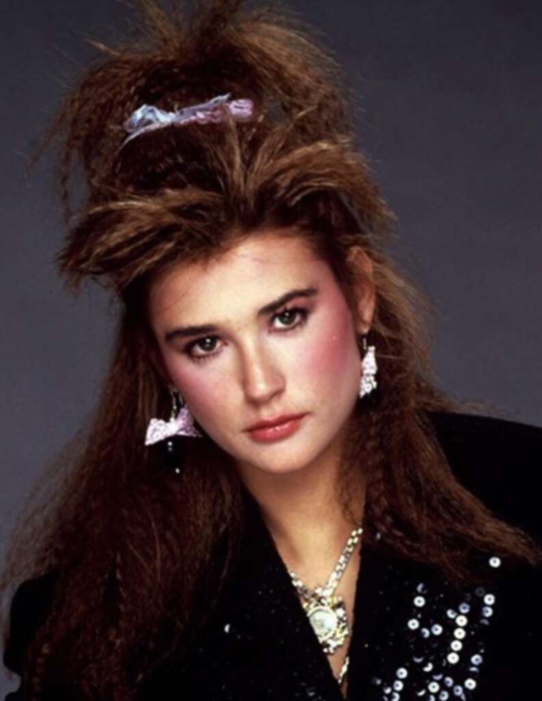 A woman with voluminous, teased brown hair adorned with a light purple scrunchie and large pink butterfly earrings. She is wearing a black sequined outfit and has pinkish makeup, standing against a plain dark background.