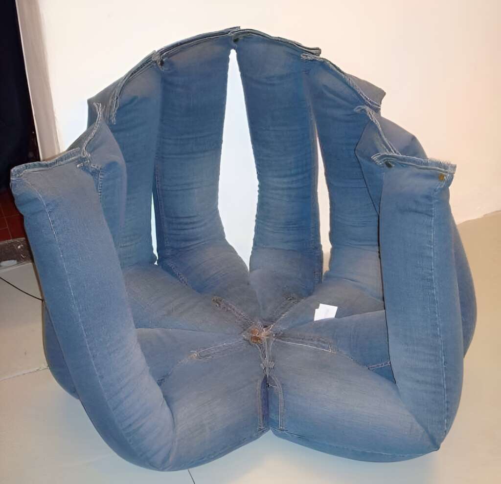 A unique chair made from blue denim jeans stitched together, forming a cushioned circular seat with multiple vertical sections resembling the legs and waistband of the jeans. The chair sits on a beige floor against a white wall.