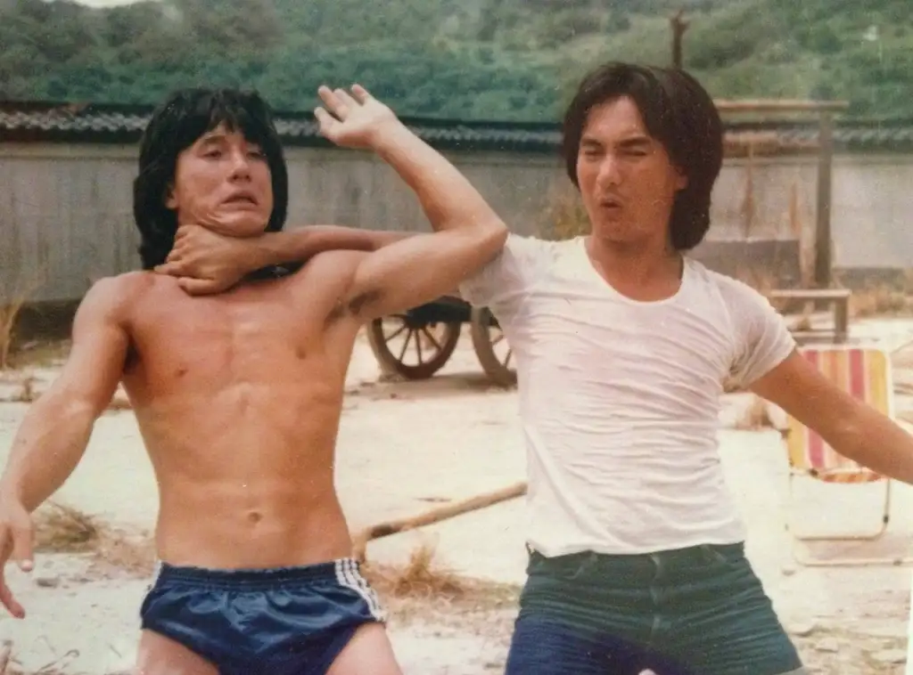 Two men are playfully mock-fighting outdoors. The man on the left, shirtless in blue shorts, is grabbing the neck of the man on the right, who is wearing a white T-shirt and jeans. Both have expressive faces, and a rustic building and cart are visible in the background.