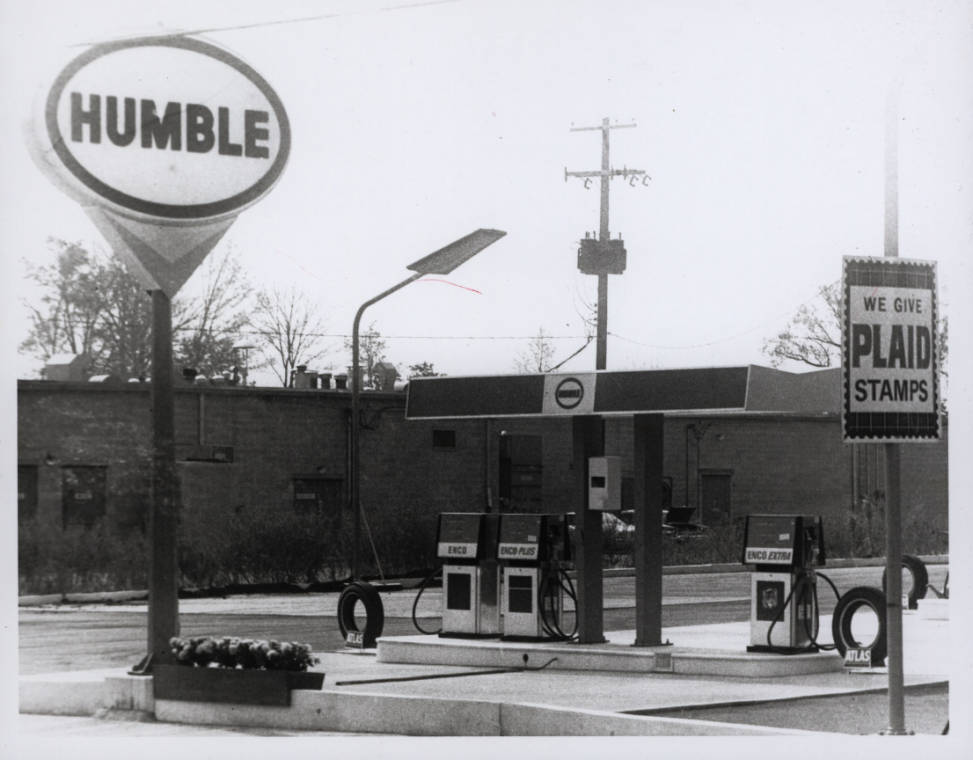 A black-and-white image of a vintage gas station. A large sign with "Humble" is prominently displayed on the left. The gas pumps are in the center, with smaller signs and a phone booth next to them, and a sign on the right saying, "We Give Plaid Stamps.