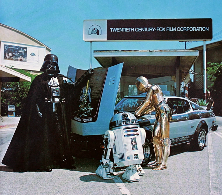 Darth Vader, R2-D2, and C-3PO stand by a car with its hood up, revealing a Star Wars graphic. They are in front of the Twentieth Century-Fox Film Corporation building.