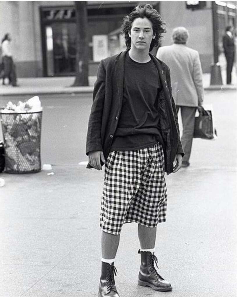 A young man with long hair stands on a city street wearing a dark jacket over a t-shirt, checkered shorts, and boots. A trash bin and a person walking away carrying a briefcase are visible in the background. The image is black and white.