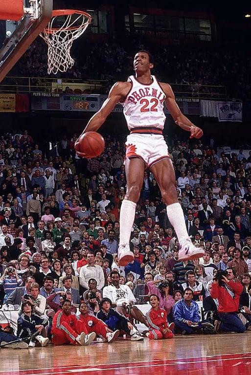 A basketball player, wearing a white jersey with the number 22 and "Phoenix" on it, is mid-air, seemingly in the middle of a slam dunk. The court is surrounded by a large, enthusiastic crowd. Several people, including other players, are seated on the sidelines.