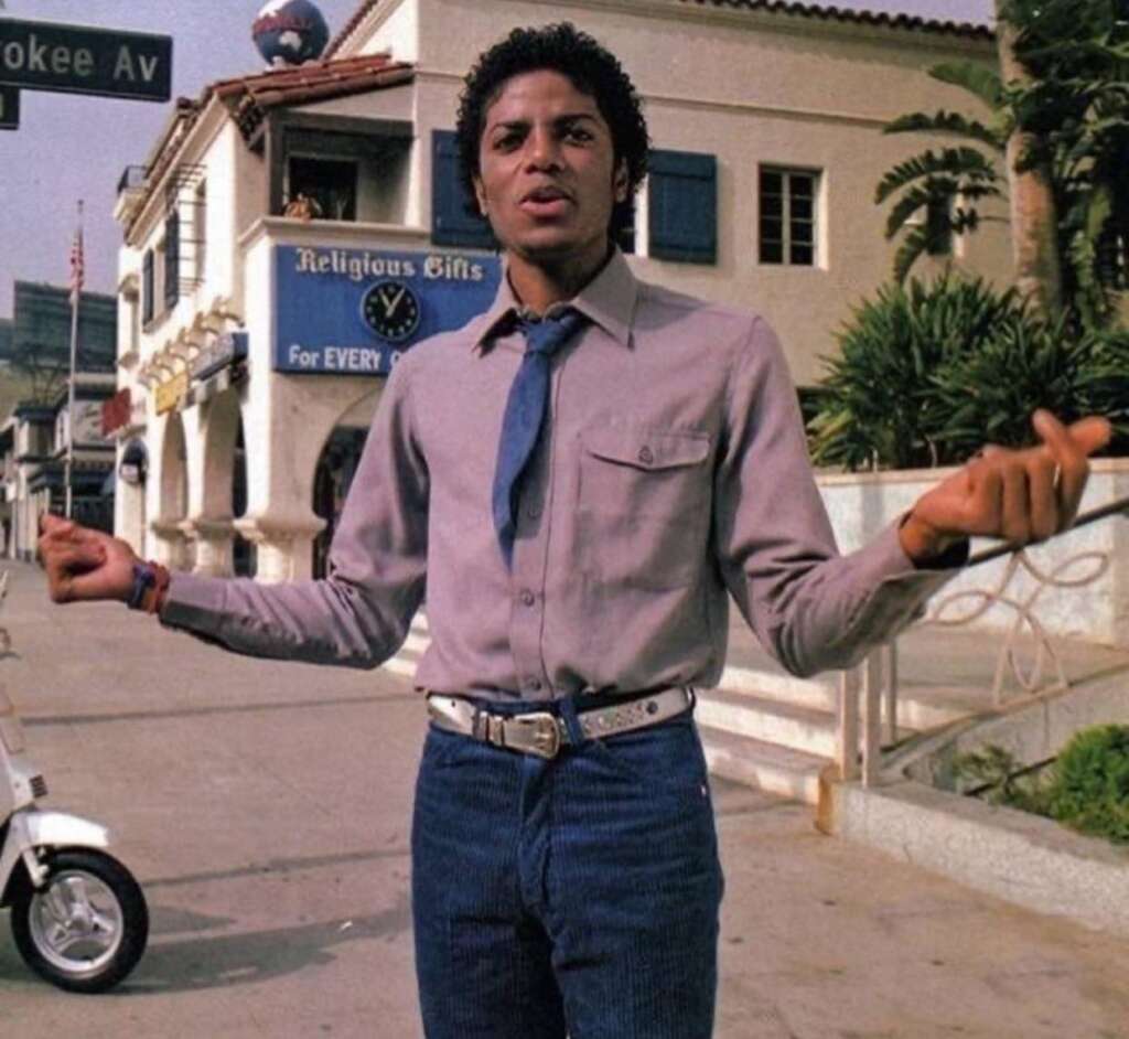 A person wearing a light purple shirt, dark blue jeans, and a blue tie stands on the sidewalk with arms outstretched. In the background, there's a building with a sign reading "Religious Gifts" and a parked scooter.