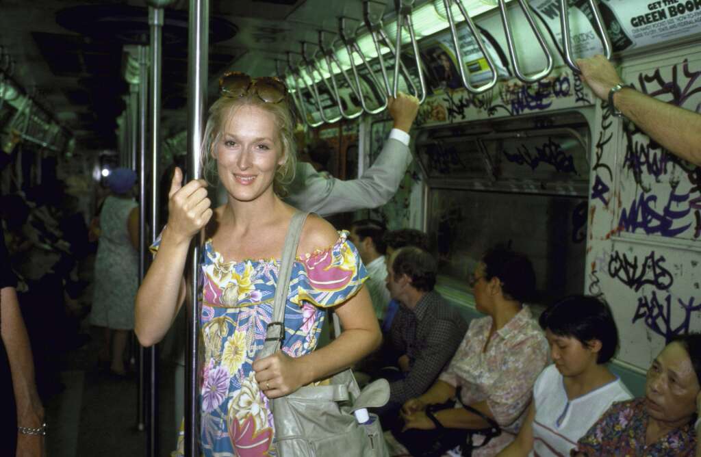 A woman stands in a subway car holding a pole with one hand. She wears a colorful floral dress and carries a bag over her shoulder. Other passengers sit around, and graffiti covers the walls of the subway car. The lighting is dim, and the atmosphere appears casual.