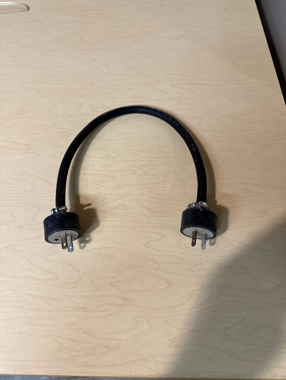 A black U-shaped power cable with plug prongs at both ends lies on a light wooden surface. The cable has metal and plastic connectors at each end, and appears to be an adapter or connector of some sort for electrical purposes.