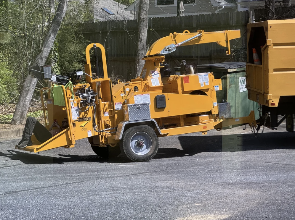 A yellow wood chipper is parked on the side of the road, attached to a wood disposal container. It has safety labels, controls, and a chute for processing branches. Trees and a wooden fence are visible in the background.