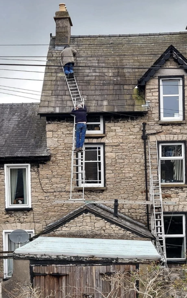 Two people scaling a slate roof, using ladders. The upper person is near the chimney, holding a wire, while the lower person stabilizes the ladder from below. The old stone house has multiple windows and visible roof details, such as moss and weathering.