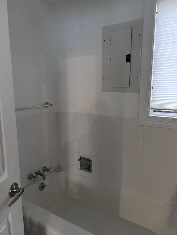 A bathroom with a white tile bathtub featuring a faucet and a showerhead mount. A towel bar is installed above the tub. There's a rectangular electrical panel on the wall above the tub. A window with closed blinds is visible on the right side of the image.