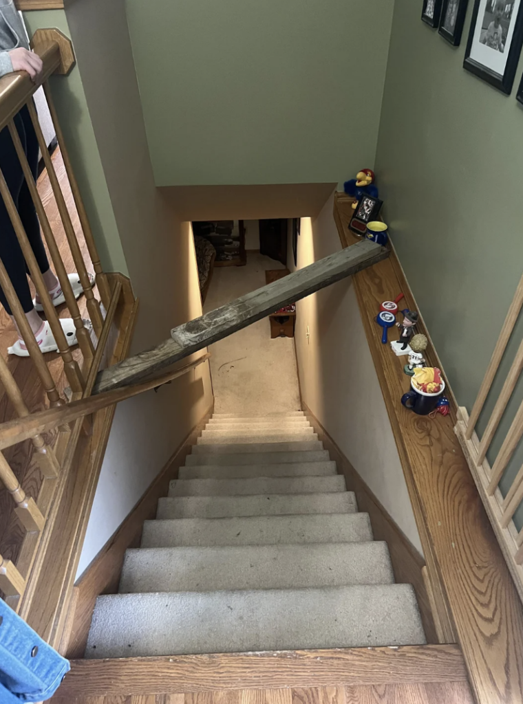 A wooden plank is precariously balanced across a staircase, forming a bridge from the top step to the railing. Various knick-knacks line the railing. Two people stand at the bottom of the plank. Caution is implied due to the setup.