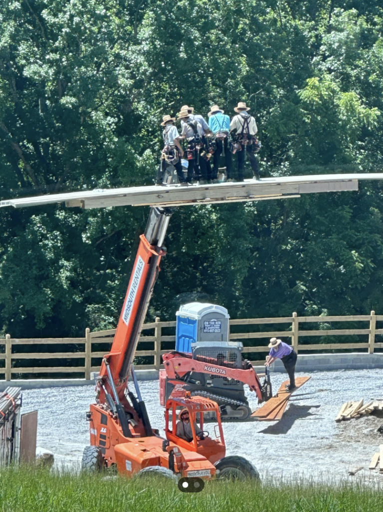 A group of construction workers standing on a wooden plank elevated by a large forklift, with a portable toilet and another worker on the ground in the background. Trees and a wooden fence are visible, and the scene takes place on a gravel road.