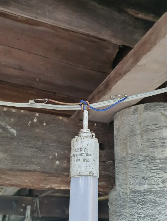 A fluorescent LED tube light is hanging from a wooden ceiling. The light is connected to an electrical wire using metal wire clips. The background shows wooden beams and an unfinished ceiling structure.