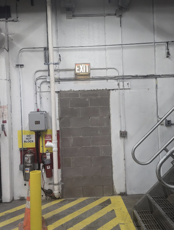 The image shows a wall with a bricked-up door where an illuminated exit sign hangs above it. Beside the door, there are fire extinguishers, a fire hose, and a "Do Not Block" sign. Nearby are a metal staircase and a yellow-striped floor.