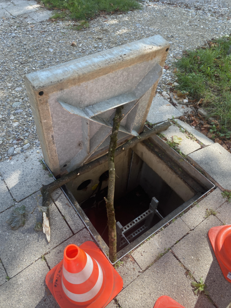 An open manhole with a metal cover propped up by a stick. There are orange traffic cones placed around the manhole, and a ladder leading down into the hole. The surrounding area has grass and paved pathways.