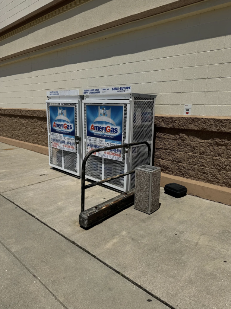 A propane tank exchange area outdoors next to a building. There are two metal cages labeled "AmeriGas" holding propane tanks. A metal bike rack and a rectangular trash can are nearby on the concrete ground.