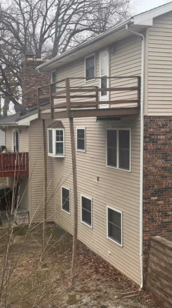 A beige house has a second-floor balcony held up by a single, long, angled wooden support post. The balcony appears unsafe, with the support post looking unstable. There are bare trees in the background and other house structures visible.