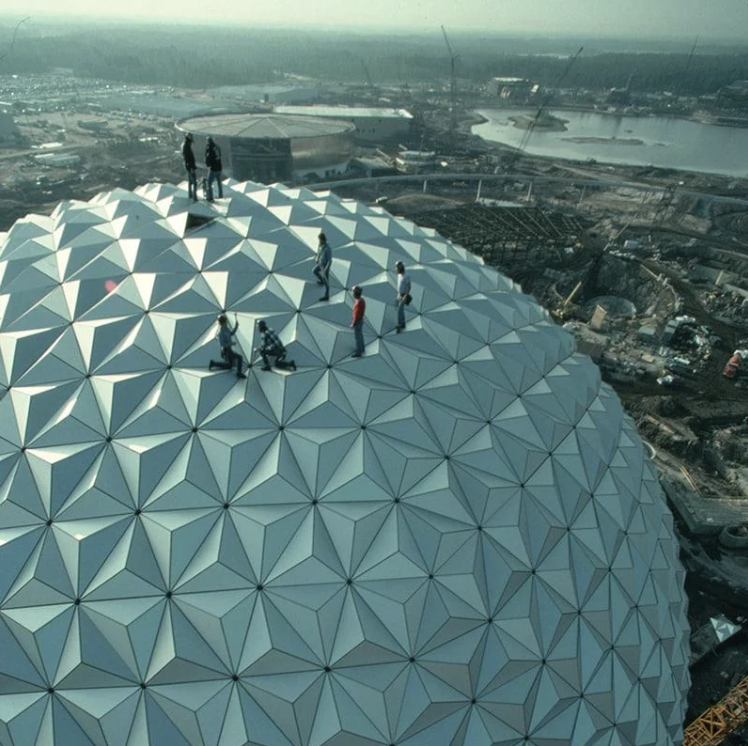 A group of people stands and sits atop the geometric metal surface of a large, dome-shaped structure, possibly under construction. The background reveals a sprawling construction site with cranes, buildings, and a body of water under a hazy sky.