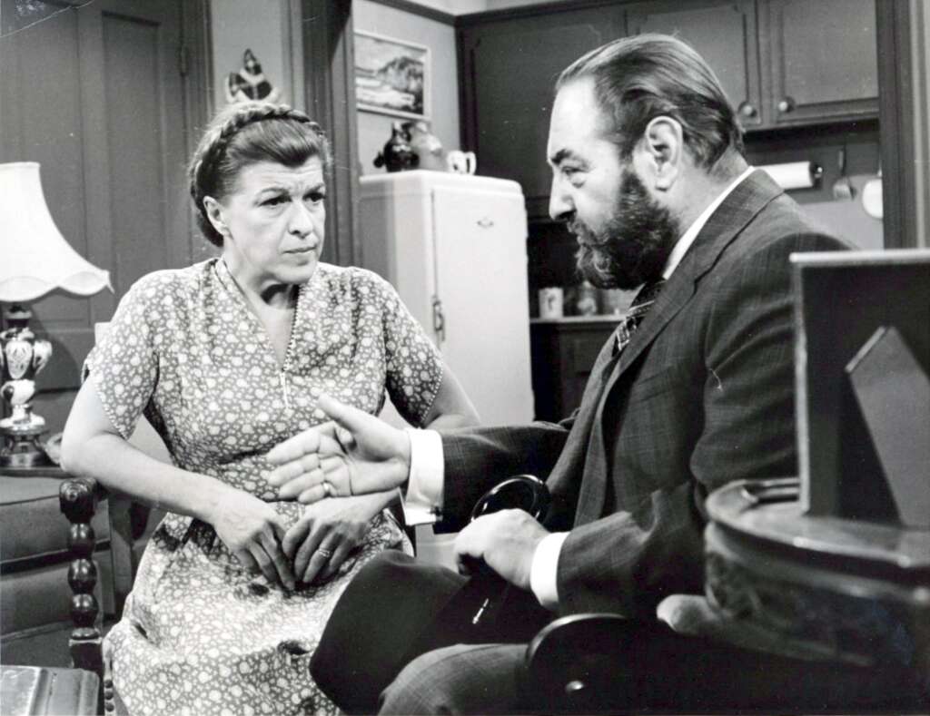 A woman in a floral dress sits on a sofa looking critically at a bearded man in a suit who is gesturing with his hand. They are in a modestly decorated room with a lamp, a refrigerator, and various items on a table and shelves in the background.
