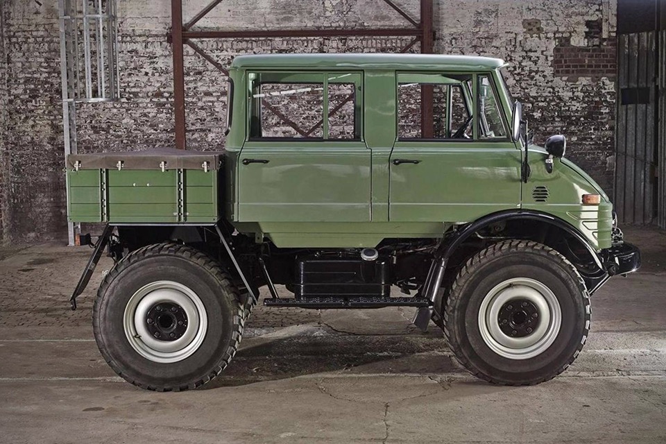 A green, vintage Unimog truck with a double cab is parked indoors against a distressed brick wall. The truck has a sturdy, industrial build, large off-road tires, and a flatbed cargo area. The setting appears to be an old warehouse or garage.