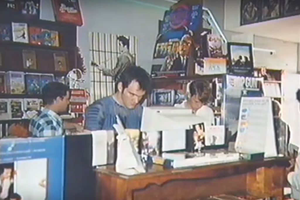 Three people are seen working behind the counter of a busy record store. The shop is filled with vinyl records and posters on the walls. The individuals appear focused on their tasks amid a backdrop of musical paraphernalia. A large poster and a computer are notable on the counter.