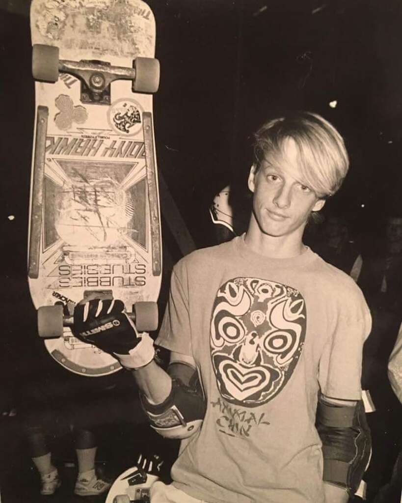 A black-and-white photo shows a young person with skateboard gear, holding up a skateboard vertically. The skateboard features various stickers. The person is wearing a t-shirt with a large mask-like design on it and protective wrist guards.