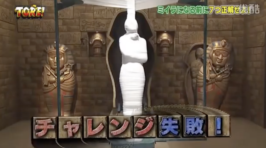 A scene from a game show features a fully wrapped mummy figure standing upright in a glass case. It is flanked by statues in niches with hieroglyphs on the walls. A caption in Japanese reads: "チャレンジ失敗!" (Challenge failed!).