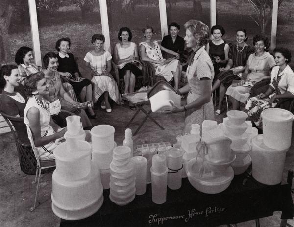 A group of women seated in a circle indoors while one woman stands and demonstrates the use of plastic containers. Various Tupperware items are displayed on a table in front of her. The scene appears to be a Tupperware home party from the mid-20th century.