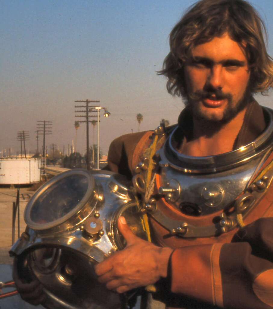 A person with long hair and a beard is holding an old-fashioned diving helmet, standing outdoors. The individual is dressed in a brown diver's suit. In the background, there are electrical poles, palm trees, and an industrial setting under a clear sky.