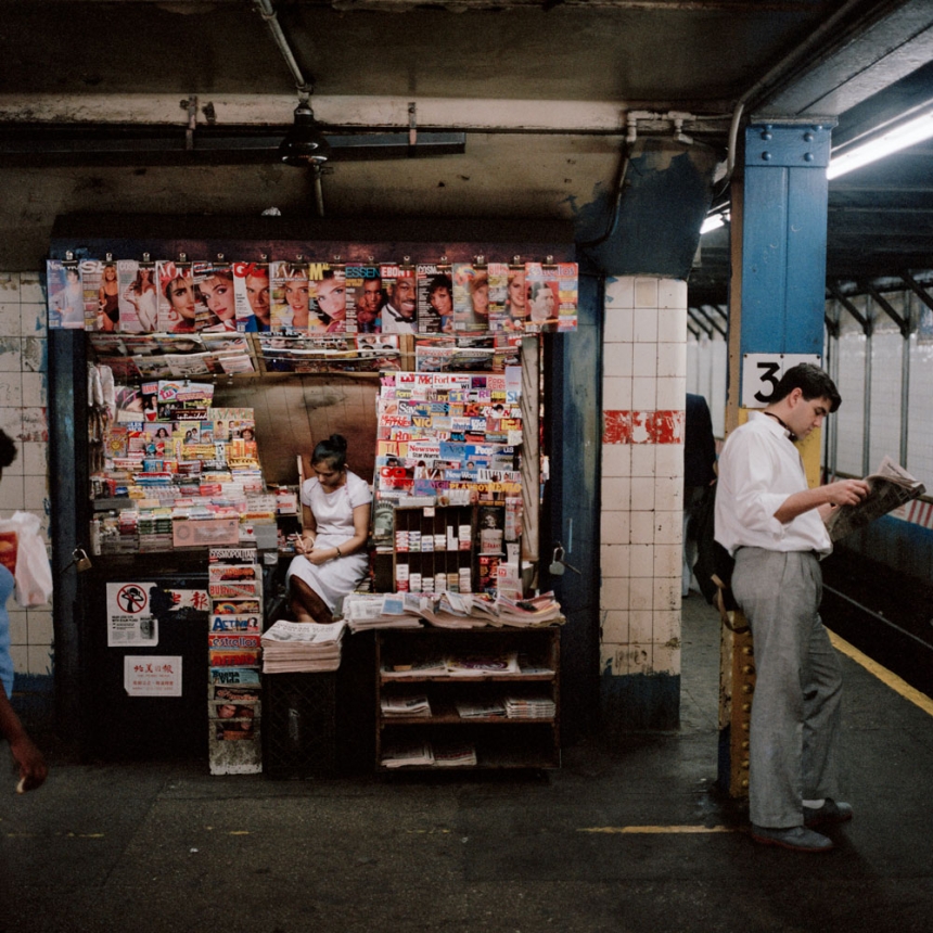 A newsstand in a subway station offers a variety of magazines and newspapers. The vendor is seated inside, while a man stands nearby, reading a magazine. Another individual is walking past the newsstand. The station appears to be slightly worn with visible tile walls.