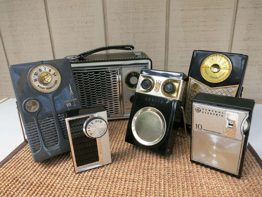 A collection of vintage transistor radios is displayed on a woven mat, set against a wooden backdrop. The radios vary in design, featuring circular dials, grids, and different brand names such as Zenith and General Electric. The assortment showcases styles from a bygone era.