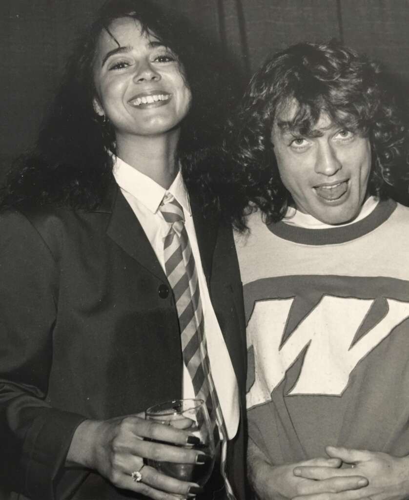 A black and white photo of two people smiling. The person on the left is wearing a suit with a tie and holding a drink. The person on the right is wearing a shirt with a large "W" on it and making a playful expression.