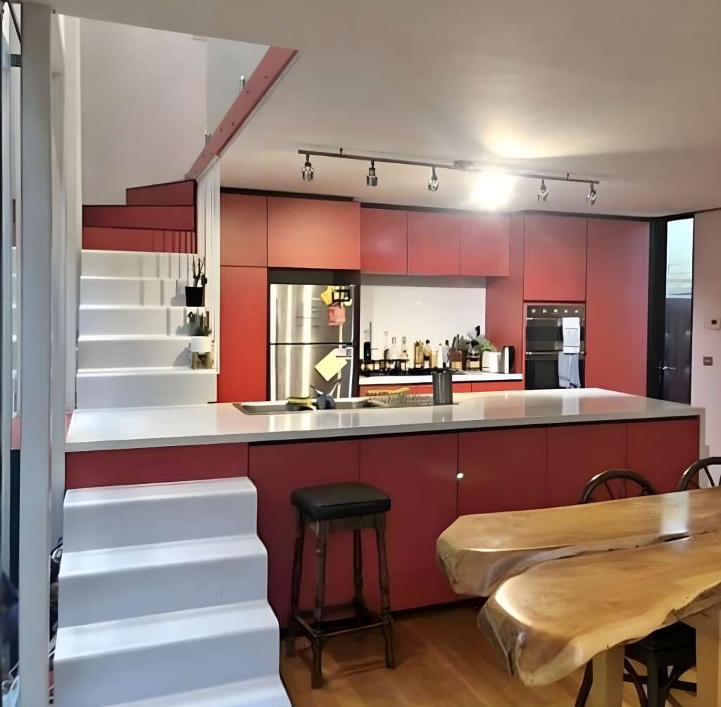 A modern kitchen with red cabinetry, a white countertop, and built-in stainless steel appliances. Behind the counter is a well-stocked bar area. A wooden dining table with chairs is in the foreground, and a staircase with white steps is on the left.