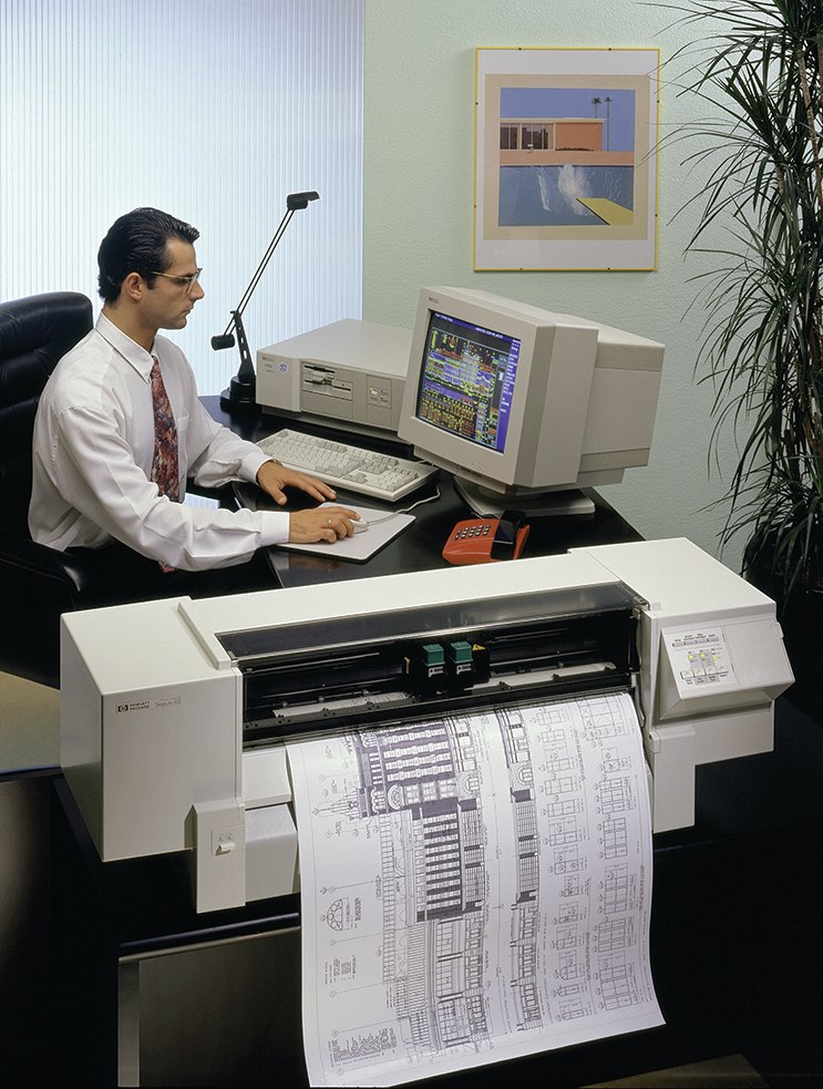 A man in formal attire is working at a desk with a vintage computer and a large printer. The printer is producing technical architectural drawings. A framed architectural artwork hangs on the wall in the background. The workspace is well-organized and professional.