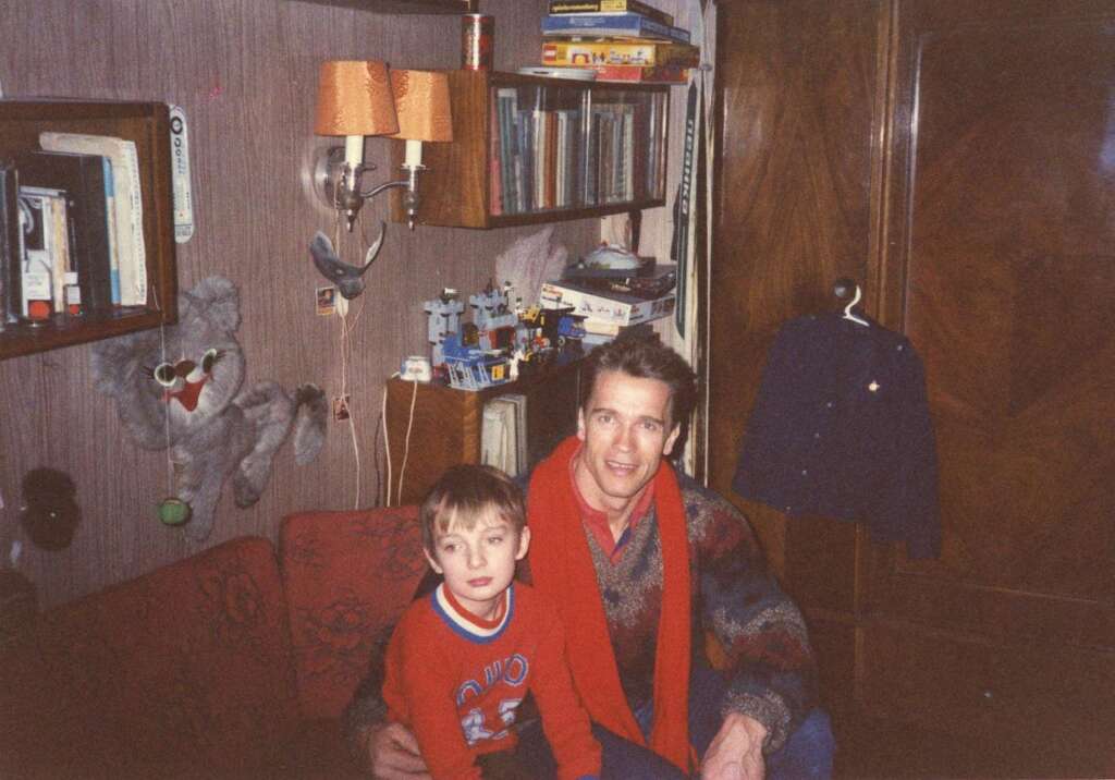 A man and a child sit together on a red couch in a room filled with books, toys, and shelves. The man is wearing a sweater and a red scarf, and the child is dressed in a red sweater with a blue print. The background features various items including a wall-mounted lamp.