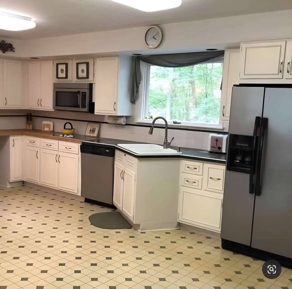 A spacious kitchen featuring white cabinets, a stainless steel refrigerator, microwave, dishwasher, and a black countertop. There's a large window above the sink with a view of greenery outside. The floor has a beige and green patterned tile design. A clock hangs above the window.