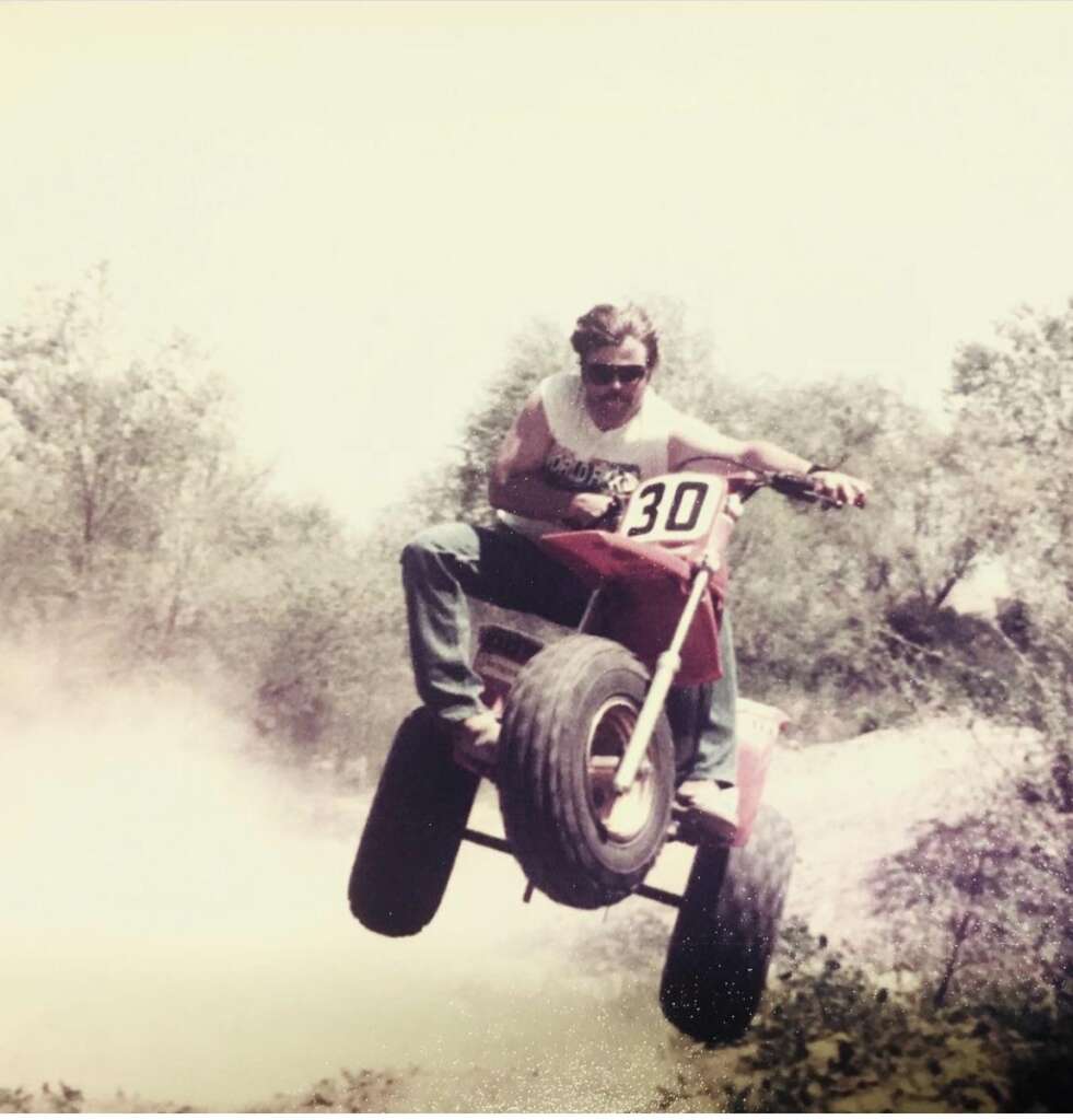 A person wearing sunglasses and a sleeveless shirt rides a red ATV with the number 30 in the air, kicking up dust. The background shows trees and bushes under a clear sky.