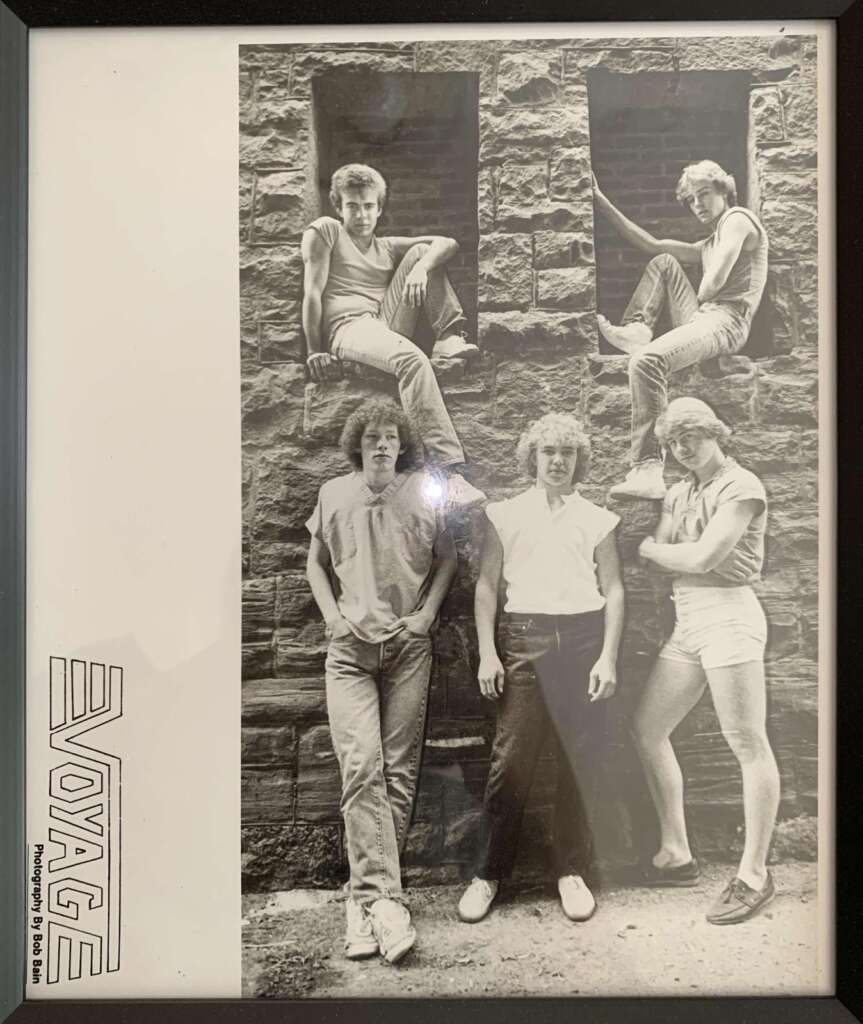 A black-and-white photo of five young men posing against a stone wall with two small windows. The text "VOYAGE" and "Photograph by Rob Lees" is printed on the left side. Some members are standing while others sit or lean on the wall.