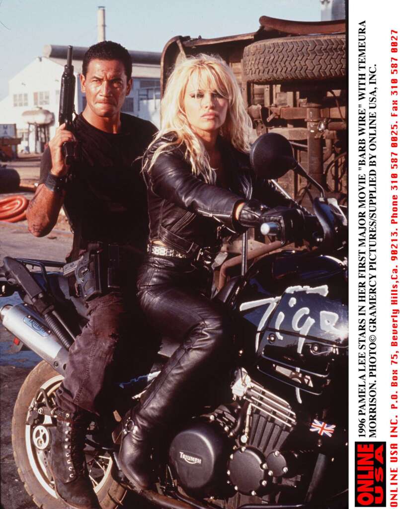 A man in black clothing and a woman with blonde hair in a leather outfit pose on a motorcycle in an industrial setting. The man holds a gun, and a large tire is visible at the top of the image. Text with red accents is on the right side of the photo.