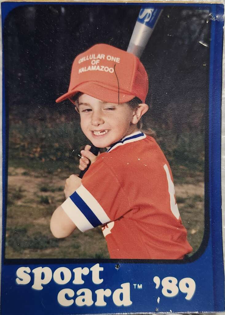 A young boy in a baseball uniform holds a bat over his shoulder, smiling with a wink. He wears a red cap with white text that reads "CELLULAR ONE OF KALAMAZOO" and a red and white jersey. The baseball card is from 1989 and labeled "sport card '89".