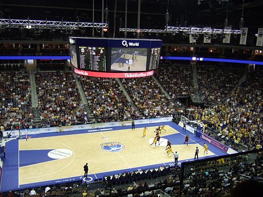 A professional basketball game is being played in a large indoor arena with a full crowd. The scoreboard shows game statistics, and the court is brightly lit. The venue features several advertisement banners, and spectators are closely watching the action.