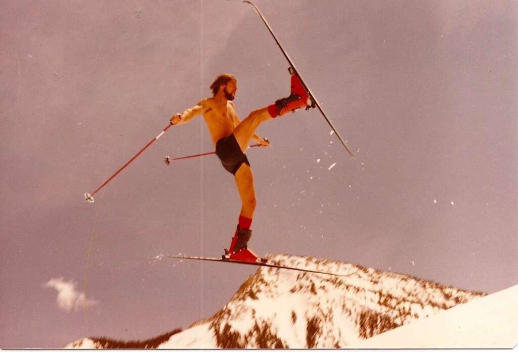 A skier in mid-air performing a trick. The skier is shirtless, wearing black shorts, red socks, and red ski boots. They are holding ski poles and their skis are crossed. There is a snowy mountain with a treeline in the background under a clear sky.