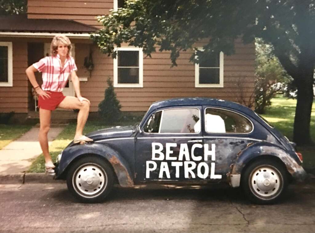 A person with shoulder-length blond hair in a red and white striped shirt and red shorts stands with one leg on the front bumper of an old blue Volkswagen Beetle with the words "BEACH PATROL" painted on the door. A brown house and greenery are in the background.