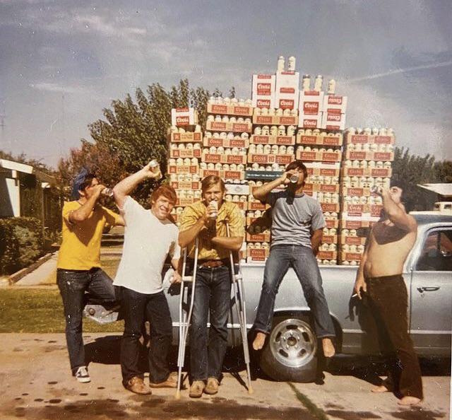 A group of five men are gathered in front of a pickup truck with a large stack of beer boxes in its bed. Four of the men are holding beer cans, and one is on crutches. They appear to be in a residential area with trees and houses in the background.