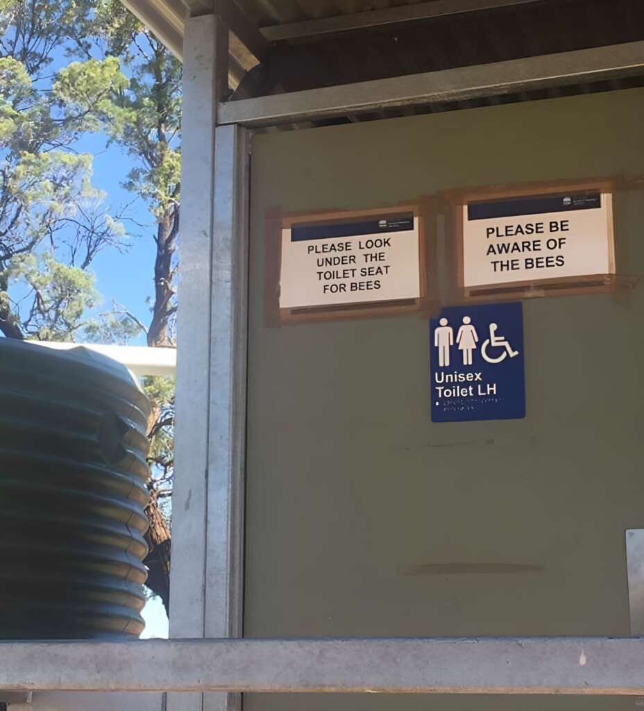 A public restroom with a sign showing it is a unisex toilet. Two additional signs above warning users to "Please look under the toilet seat for bees" and "Please be aware of the bees". The restroom is outdoors, surrounded by trees and a water tank is visible nearby.